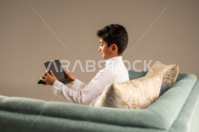 استخدام الأجهزة التقنية الحديثة، بورتريه مقرب من الخلف لطفل عربي سعودي خليجي يرتدي الثوب التقليدي يجلس على كرسي مريح ويقوم باستخدام التابلت، مفهوم التعليم وإنجاز المهام عن بعد، قضاء وقت ممتع على الإنترنت، اللعب على الجهاز اللوحي، خلفية بيج