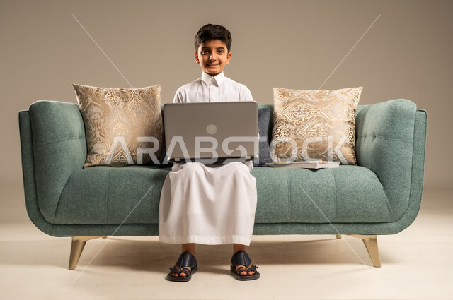 The concept of distance learning using modern technical devices and technology, following lessons online, a portrait of a Saudi Arabian Gulf child wearing traditional clothing looking at the camera and sitting on a comfortable sofa in front of a laptop, beige background