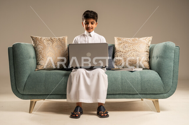 مفهوم التعلم عن بعد باستخدام الأجهزة التقنية الحديثة والتكنولوجيا، متابعة الدروس أونلاين، بورتريه لطفل عربي خليجي سعودي يرتدي الزي التقليدي يجلس على اريكة مريحة ينظر الى الحاسوب المحمول , خلفية بيج
