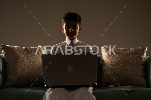 متابعة الدروس أونلاين، مفهوم التعلم عن بعد باستخدام الأجهزة التقنية الحديثة والتكنولوجيا، بورتريه ظليلي مقرب لطفل عربي خليجي سعودي يرتدي الزي التقليدي ينظر الى الكاميرا يجلس على اريكة مريحة امام الحاسوب المحمول , خلفية بيج
