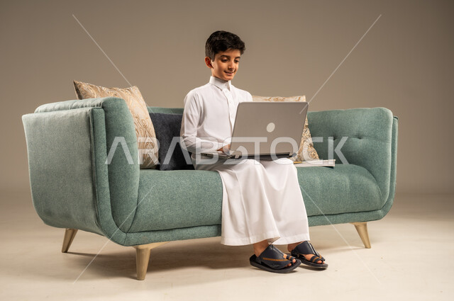متابعة الدروس أونلاين، مفهوم التعلم عن بعد باستخدام الأجهزة التقنية الحديثة والتكنولوجيا، بورتريه لطفل عربي خليجي سعودي يرتدي الزي التقليدي يجلس على اريكة مريحة ينظر الى الحاسوب المحمول , خلفية بيج