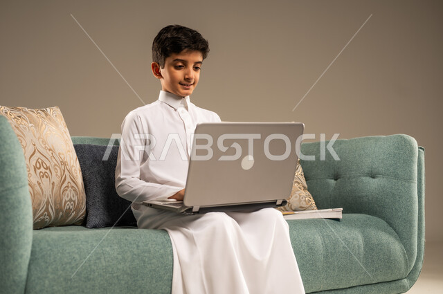 متابعة الدروس أونلاين، مفهوم التعلم عن بعد باستخدام الأجهزة التقنية الحديثة والتكنولوجيا، بورتريه مقرب لطفل عربي خليجي سعودي يرتدي الزي التقليدي يجلس على اريكة مريحة ينظر الى الحاسوب المحمول , خلفية بيج