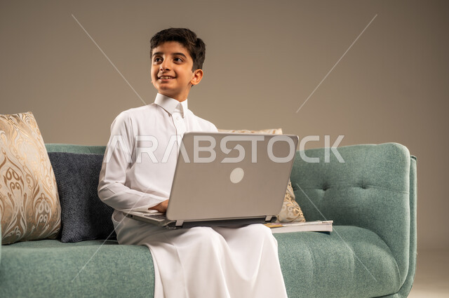 متابعة الدروس أونلاين، مفهوم التعلم عن بعد باستخدام الأجهزة التقنية الحديثة والتكنولوجيا، بورتريه مقرب لطفل عربي خليجي سعودي يرتدي الزي التقليدي يجلس على اريكة مريحة أمام الحاسوب المحمول ينظر الى شيء ما, خلفية بيج