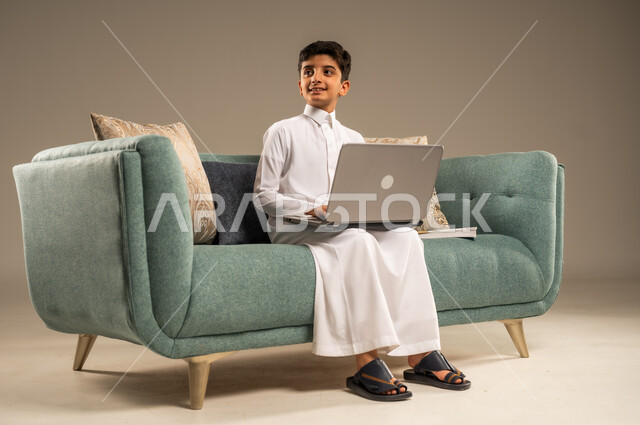 متابعة الدروس أونلاين، مفهوم التعلم عن بعد باستخدام الأجهزة التقنية الحديثة والتكنولوجيا، بورتريه لطفل عربي خليجي سعودي يرتدي الزي التقليدي يجلس على اريكة مريحة أمام الحاسوب المحمول ينظر الى شيء ما, خلفية بيج
