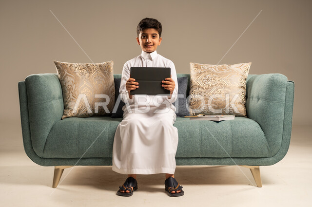  التسليه والترفيه على الأجهزة المحمولة, مفهوم التعليم عن بعد, بورتريه لطفل عربي سعودي خليجي يرتدي العباءة يجلس على أريكه مريحه يمسك التابليت ينظر الى الكاميرا بايماءات وجه تدل على الاستمتاع والسعادة, خلفية بيج 
