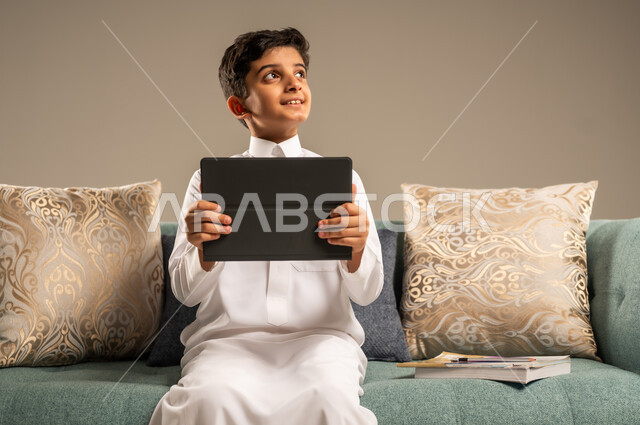 مفهوم التعليم عن بعد, بورتريه مقرب لطفل عربي سعودي خليجي يرتدي العباءة يجلس على أريكه مريحه يمسك التابليت ينظر الى شيء ما بايماءات وجه تدل على الاستمتاع والسعادة, التسليه والترفيه على الأجهزة المحمولة, خلفية بيج 