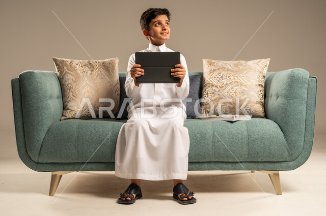 Distance learning concept, portrait of a Saudi Arabian Gulf child wearing an abaya sitting on a comfortable sofa holding a tablet looking at something with facial expressions indicating enjoyment and happiness, entertainment and amusement on mobile devices, beige background