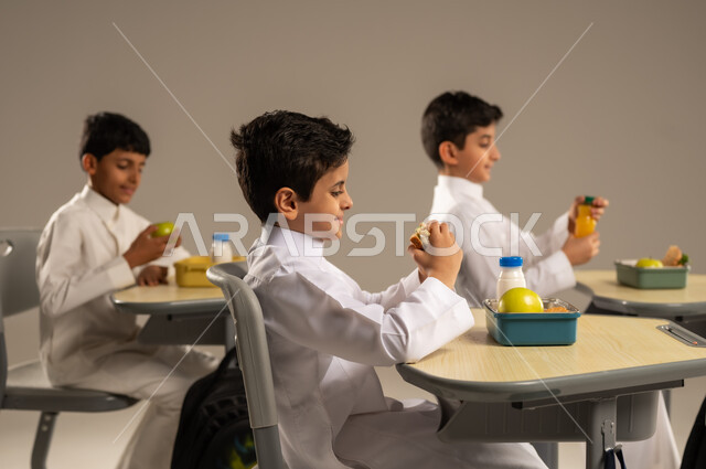 الوجبات الغنية بالعناصر الغذائية الهامة لبناء الجسم، الاستمتاع بتناول الأطعمة الصحية في وقت الفسحة المدرسية، موسم العودة للمدارس، بورتريه مقرب لمجموعة من الطلاب العربيين الخليجيين السعوديين يرتدون الزي المدرسي يجلسون في مقاعدهم وأمامهم اللانش البوكس، خلفية بيج