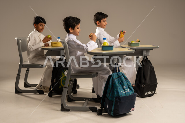 الوجبات الغنية بالعناصر الغذائية الهامة لبناء الجسم، الاستمتاع بتناول الأطعمة الصحية في وقت الفسحة المدرسية، موسم العودة للمدارس، بورتريه لمجموعة من الطلاب العربيين الخليجيين السعوديين يرتدون الزي المدرسي يجلسون في مقاعدهم وأمامهم اللانش البوكس، خلفية بيج