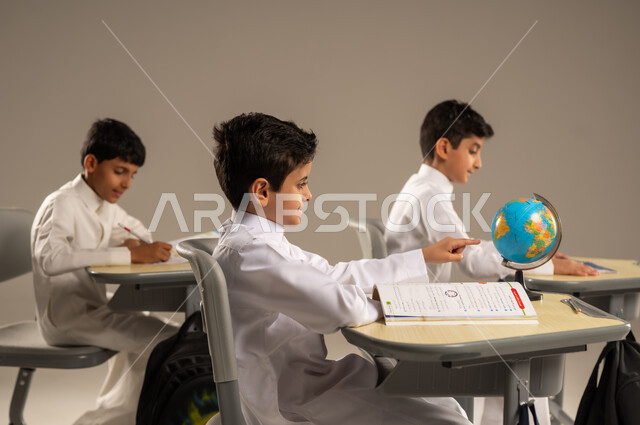 The concept of exploration and research around the world, the development of teaching methods in the Kingdom's schools, the quality of effective learning methods and the use of tools and aids, a portrait of Saudi Arabian Gulf students wearing school uniforms sitting in their seats with a miniature globe in front of them, beige background