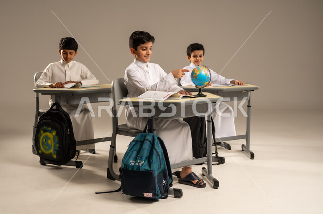 The development of teaching methods in Saudi schools, the concept of exploration and research around the world, the quality of effective learning methods and the use of tools and aids, a portrait of Saudi Gulf Arab students wearing school uniforms sitting in their seats with a miniature globe in front of them, beige background