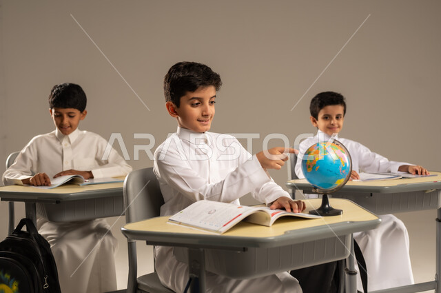 تطور طرائق التدريس في مدارس السعودية، مفهوم الاستكشاف والبحث حول العالم، جودة أساليب التعلم الفعال وباستخدام الأدوات والوسائل المساعدة، بورتريه مقرب لطلاب عربيين خليجيين سعوديين يرتدون الزي المدرسي يجلسون في مقاعدهم وأمامهم مجسم مصغر للكرة الأرضية، خلفية بيج