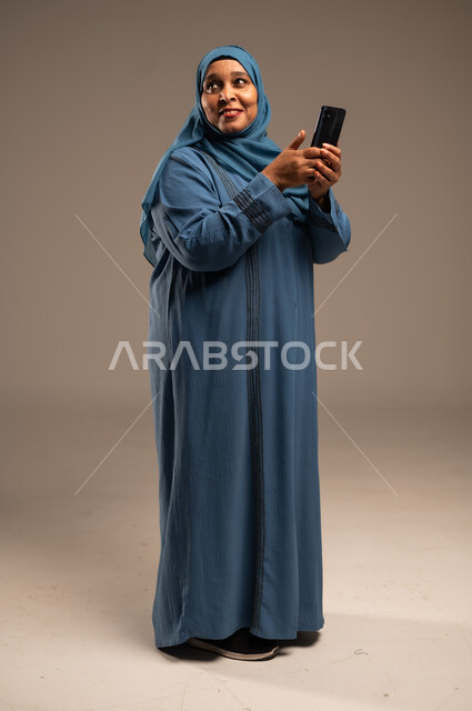 Browsing social media applications and programs, exchanging conversations with family and friends, portrait of an elderly veiled Saudi Arabian Gulf woman wearing an abaya holding a mobile phone in her hand, looking at something, using modern and advanced devices and technologies, full-length body, beige background