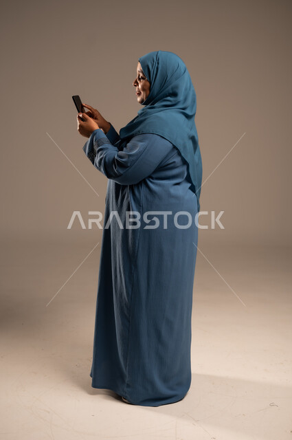Browsing social media applications and programs, exchanging conversations and news with family and friends, side portrait of an elderly Saudi Arabian Gulf woman wearing a hijab, holding a mobile phone in her hand, using modern and advanced devices and technologies, full-length body, beige background