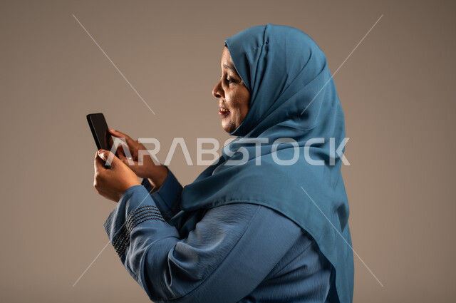 تصفح التطبيقات وبرامج التواصل الاجتماعي، تبادل الأحاديث والأخبار مع الأهل والأصدقاء، بورتريه مقرب من الجانب لإمرأة كبيرة بالسن عربية خليجية سعودية محجبة ترتدي العباءة تمسك بيدها الهاتف المحمول، استخدام الأجهزة والتقنيات الحديثة المتطورة، خلفية بيج