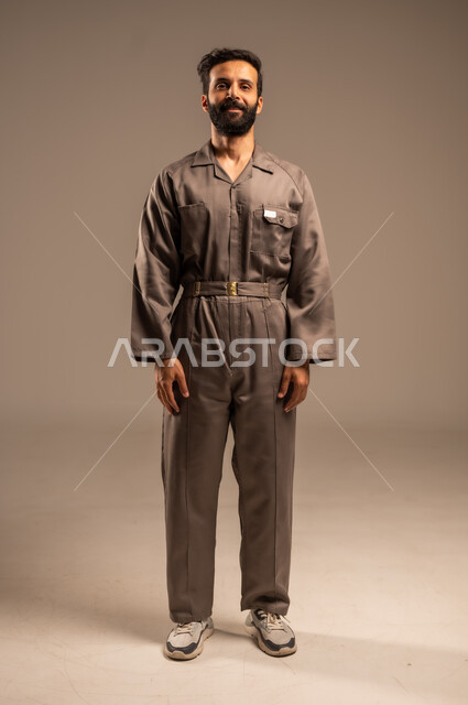  أعمال الإصلاح وورشات الصيانة، بورتريه لعامل عربي خليجي سعودي مبتسم يرتدي اليونيفورم الخاص بالعمل يقف باستقامة ينظر الى الكاميرا بتعابير السرور الثقة بالنفس, العمل في المجال الميكانيكي والكهربائي، مهن ووظائف سعودية شبابية, صورة الجسم بالطول الكامل, خلفية رمادية