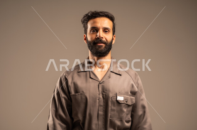  أعمال الإصلاح وورشات الصيانة، بورتريه مقرب لعامل عربي خليجي سعودي مبتسم يرتدي اليونيفورم الخاص بالعمل يقف باستقامة ينظر الى الكاميرا بتعابير السرور الثقة بالنفس, العمل في المجال الميكانيكي والكهربائي، مهن ووظائف سعودية شبابية, خلفية رمادية