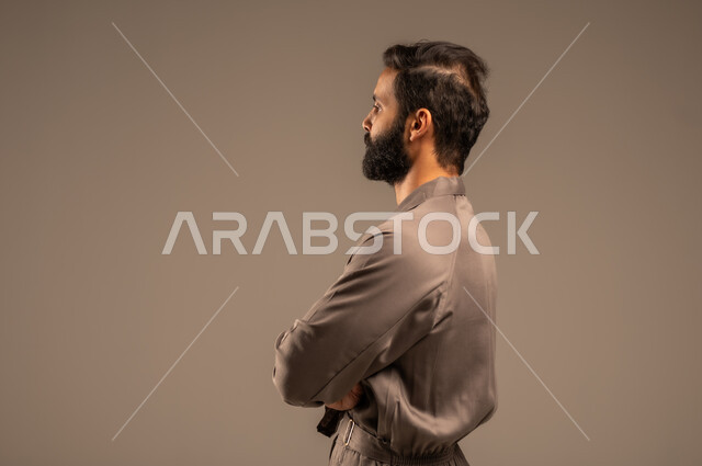 العمل في المجال الميكانيكي والكهربائي، مهن ووظائف سعودية شبابية, أعمال الإصلاح وورشات الصيانة، بورتريه مقرب من الجانب لعامل عربي خليجي سعودي يرتدي اليونيفورم الخاص بالعمل يقف بإيدي متقاطعة بتعابير الثقة بالنفس, خلفية رمادية