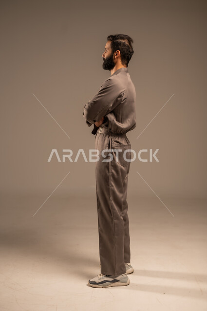 العمل في المجال الميكانيكي والكهربائي، مهن ووظائف سعودية شبابية, أعمال الإصلاح وورشات الصيانة، بورتريه من الجانب لعامل عربي خليجي سعودي يرتدي اليونيفورم الخاص بالعمل يقف بإيدي متقاطعة بتعابير الثقة بالنفس, الجسم بالطول الكامل, خلفية رمادية