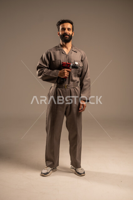 Manual measuring and inspection tools, mechanical and electrical work, repair and maintenance workshops, portrait of a Saudi Arabian Gulf worker wearing a work uniform holding an electric drill, Saudi youth professions and jobs, full body, gray background