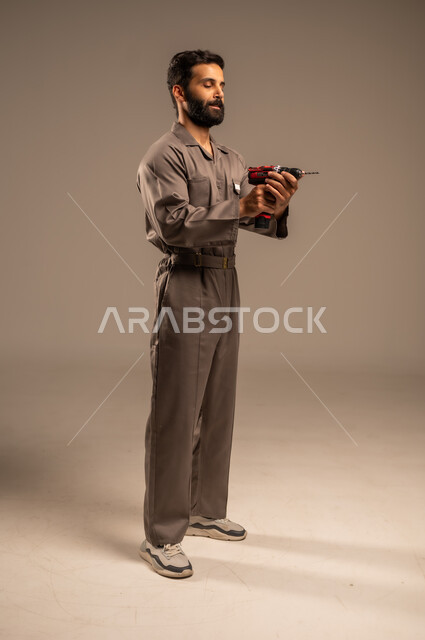 أدوات القياس والفحص اليدوية، العمل في المجال الميكانيكي والكهربائي، أعمال الإصلاح وورشات الصيانة، بورتريه لعامل عربي خليجي سعودي يرتدي اليونيفورم الخاص بالعمل يمسك بيديه جهاز التثقيب الكهربائي، مهن ووظائف سعودية شبابية، الجسم بالطول الكامل, خلفية رمادية