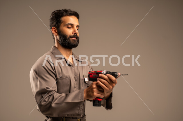 العمل في المجال الميكانيكي والكهربائي، أعمال الإصلاح وورشات الصيانة، بورتريه مقرب لعامل عربي خليجي سعودي يرتدي اليونيفورم الخاص بالعمل يمسك بيديه جهاز التثقيب الكهربائي، مهن ووظائف سعودية شبابية، أدوات القياس والفحص اليدوية، خلفية رمادية