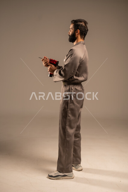 أدوات القياس والفحص اليدوية، العمل في المجال الميكانيكي والكهربائي، أعمال الإصلاح وورشات الصيانة، بورتريه لعامل عربي خليجي سعودي يرتدي اليونيفورم الخاص بالعمل يمسك بيديه جهاز التثقيب الكهربائي، مهن ووظائف سعودية شبابية، الجسم بالطول الكامل, خلفية رمادية