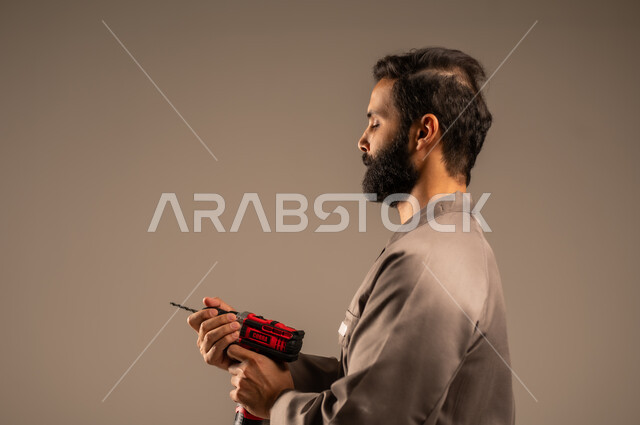العمل في المجال الميكانيكي والكهربائي، أعمال الإصلاح وورشات الصيانة، بورتريه مقرب لعامل عربي خليجي سعودي يرتدي اليونيفورم الخاص بالعمل يمسك بيديه جهاز التثقيب الكهربائي، مهن ووظائف سعودية شبابية، أدوات القياس والفحص اليدوية، خلفية رمادية