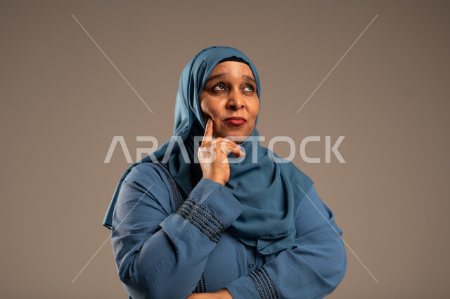 شرود الذهن والنظر الى شيء ما، التأمل والتمعن والاندماج, بورتريه مقرب لإمرأة كبيرة بالسن عربية خليجية سعودية محجبة ترتدي العباءة الزرقاء تضع يدها على خدها بإيماءات التفكير، خلفية بيج