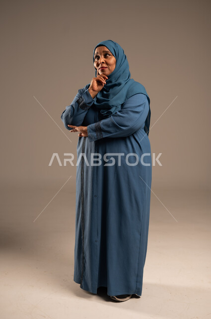شرود الذهن والنظر الى شيء ما، التأمل والتمعن والاندماج, بورتريه لإمرأة كبيرة بالسن عربية خليجية سعودية محجبة ترتدي العباءة الكحلية تضع يدها على خدها بإيماءات التفكير، الجسم بالطول الكامل, خلفية بيج