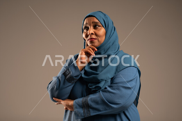 شرود الذهن والنظر الى شيء ما، التأمل والتمعن والاندماج, بورتريه مقرب من الجانب لإمرأة كبيرة بالسن عربية خليجية سعودية محجبة ترتدي العباءة الزرقاء تضع يدها على خدها بإيماءات التفكير، خلفية بيج