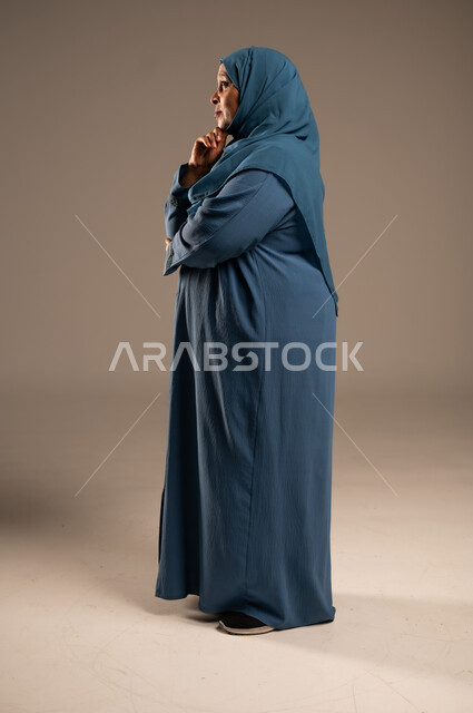 التأمل والتمعن والاندماج, شرود الذهن والنظر الى شيء ما، بورتريه من الجانب لإمرأة كبيرة بالسن عربية خليجية سعودية محجبة ترتدي العباءة الكحلية تضع يدها على خدها بإيماءات التفكير، الجسم بالطول الكامل, خلفية بيج