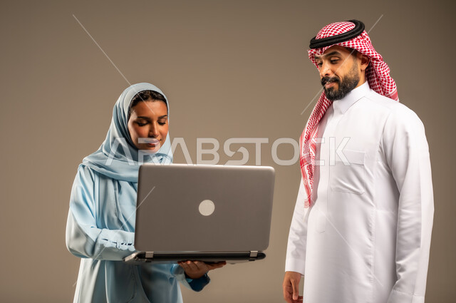 تبادل الأفكار والخبرات لتطوير استراتيجيات العمل، بورتريه مقرب لزميلين عربيين خليجيين سعوديين مبتسمين يقومان بإنجاز المهام عبر الحاسوب المحمول، التعاون وتوحيد الجهود لتحقيق أهداف الشركة، دمج التقنية و التكنولوجيا في العمل، خلفيه رمادية