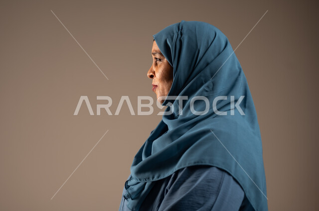 إيماءات الاعتزازو الثقة بالنفس و الانضباط، الاهتمام بالمظهر الخارجي، بورتريه مقرب من الجانب لإمراة عربية سعودية خليجية محجبة ترتدي العباءة تقف باستقامة بتعابير ايدي متماسكة، خلفية بيج