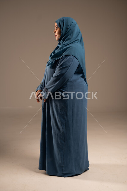 الاهتمام بالمظهر الخارجي، بورتريه من الجانب لإمراة عربية سعودية خليجية محجبة ترتدي العباءة تقف باستقامة بتعابير ايدي متماسكة، إيماءات الاعتزازو الثقة بالنفس و الانضباط، الجسم بالطول الكامل, خلفية بيج