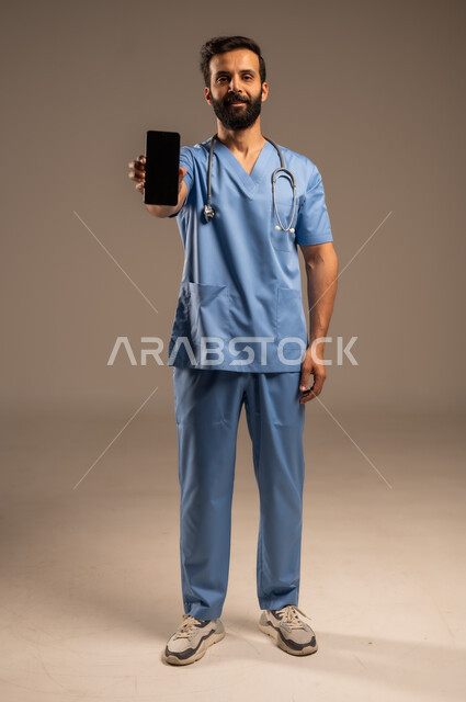 عرض التطبيقات والبرامج الصحية، بورتريه لممرض عربي خليجي سعودي يرتدي اليونيفورم الخاص بالعمل يحمل بيده الهاتف المحمول ينظر إلى الكاميرا، استخدام الأجهزة التقنية الحديثة ودمج التكنولوجيا مع الطب، شاشة سوادء فارغة، الجسم بالطول الكامل، خلفية بيج