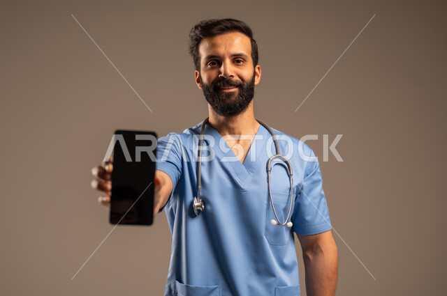 عرض التطبيقات والبرامج الصحية، بورتريه مقرب لممرض عربي خليجي سعودي يرتدي اليونيفورم الخاص بالعمل يحمل بيده الهاتف المحمول ينظر إلى الكاميرا، استخدام الأجهزة التقنية الحديثة ودمج التكنولوجيا مع الطب، شاشة سوادء فارغة، خلفية بيج