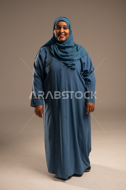 الاهتمام بالأناقة والمظهر الخارجي، إيماءات الفرح والسعادة، بورتريه لإمرأة كبيرة بالسن عربية خليجية سعودية محجبة مبتسمة ترتدي العباءة تنظر إلى الكاميرا بتعابير الاطمئنان ، التأمل والنظر إلى الأشياء السارة، الجسم بالطول الكامل, خلفية بيج