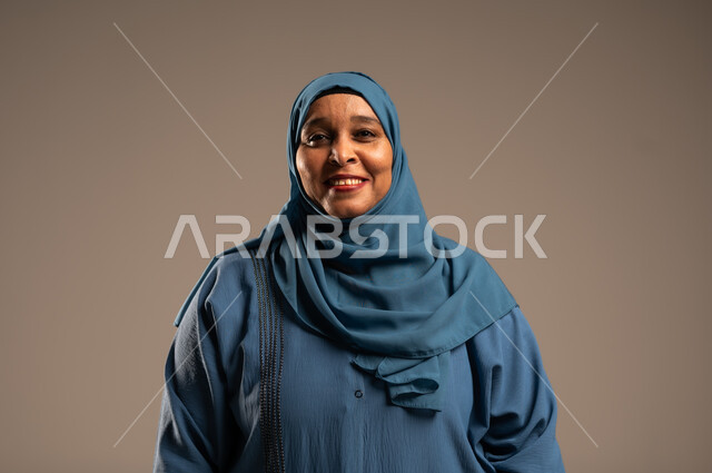 الاهتمام بالأناقة والمظهر الخارجي، إيماءات الفرح والسعادة، بورتريه مقرب لإمرأة كبيرة بالسن عربية خليجية سعودية محجبة مبتسمة ترتدي العباءة تنظر إلى الكاميرا بتعابير الاطمئنان ، التأمل والنظر إلى الأشياء السارة، خلفية بيج