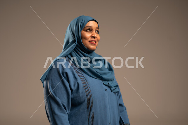 الاهتمام بالأناقة والمظهر الخارجي، إيماءات الفرح والسعادة، بورتريه مقرب من الجانب لإمرأة كبيرة بالسن عربية خليجية سعودية محجبة مبتسمة ترتدي العباءة تنظر إلى شيء ما بتعابير الاطمئنان ، التأمل بحدوث الأشياء السارة, خلفية بيج