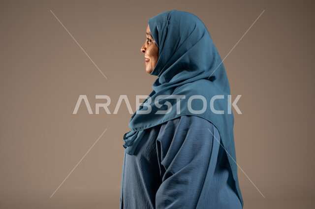 الاهتمام بالأناقة والمظهر الخارجي، إيماءات الفرح والسعادة، بورتريه مقرب من الجانب لإمرأة كبيرة بالسن عربية خليجية سعودية محجبة مبتسمة ترتدي العباءة تنظر إلى شيء ما بتعابير الاطمئنان ، التأمل بحدوث الأشياء السارة, خلفية بيج