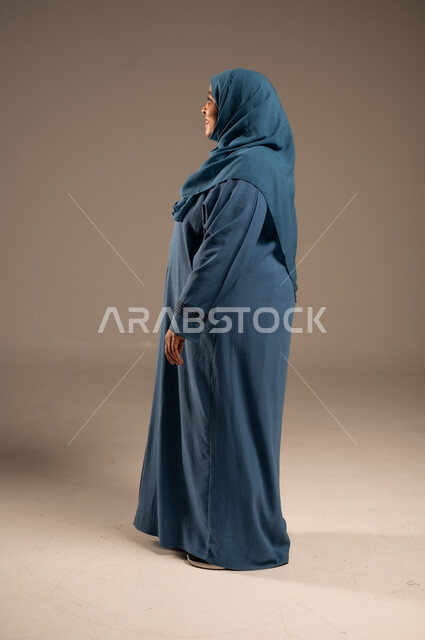 الاهتمام بالأناقة والمظهر الخارجي، إيماءات الفرح والسعادة، بورتريه من الجانب لإمرأة كبيرة بالسن عربية خليجية سعودية محجبة مبتسمة ترتدي العباءة تنظر إلى شيء ما بتعابير الاطمئنان ، التأمل بحدوث الأشياء السارة, الجسم بالطول الكامل, خلفية بيج