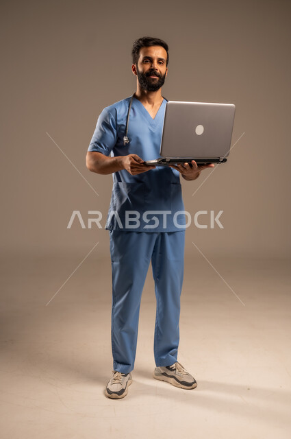 الاستعداد لتقديم الخدمات والرعاية الصحية بكل كفاءة، استخدام الأجهزة التقنية الحديثة ودمج التكنولوجيا مع الطب، بورتريه لممرض عربي خليجي سعودي يرتدي اليونيفورم الخاص بالعمل يستخدم الحاسوب المحمول ينظر إلى الكاميرا، الجسم بالطول الكامل، خلفية بيج