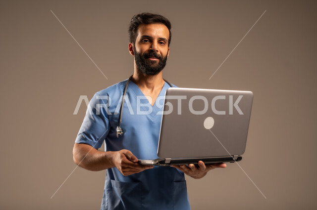 Ready to provide services and health care with all efficiency, using modern technical devices and integrating technology with medicine, close-up portrait of a Saudi Arabian Gulf nurse wearing work uniform using laptop looking at camera, beige background