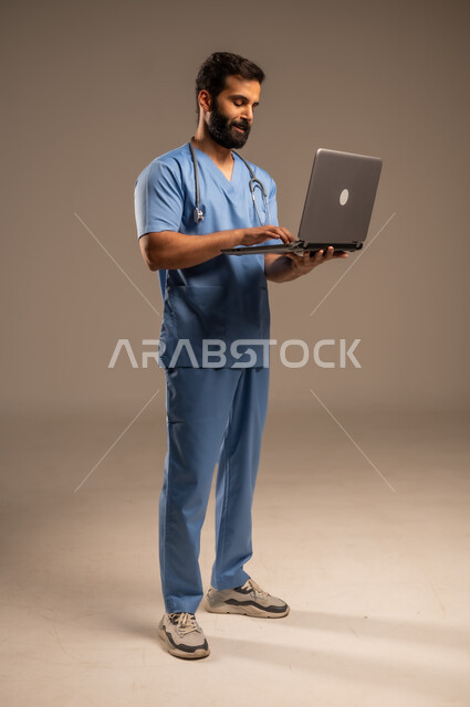 الاستعداد لتقديم الخدمات والرعاية الصحية بكل كفاءة، استخدام الأجهزة التقنية الحديثة ودمج التكنولوجيا مع الطب، بورتريه لممرض عربي خليجي سعودي يرتدي اليونيفورم الخاص بالعمل يستخدم الحاسوب المحمول، الجسم بالطول الكامل، خلفية بيج