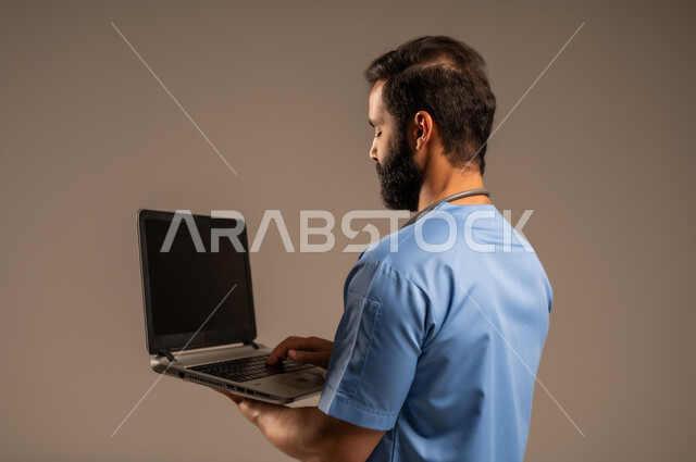 دمج التكنولوجيا مع الطب، الاستعداد لتقديم الخدمات والرعاية الصحية بكل كفاءة، بورتريه مقرب من الجانب لممرض عربي خليجي سعودي يرتدي اليونيفورم الخاص بالعمل يستخدم الحاسوب المحمول، استخدام الأجهزة التقنية الحديثة، خلفية بيج