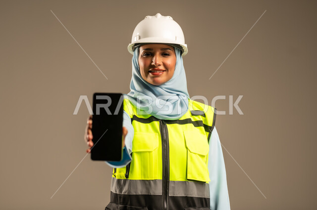 عرض شاشة سوداء فارغة عبر الهاتف المحمول, ايماءات السعادة والفرح, بورتريه مقرب لمهندسة معمارية عربية سعودية خليجية مبتسمة ترتدي الحجاب والخوذة وسترة الحماية تمسك الجوال بيدها، دمج التكنولوجيا في مجالات العمل, استخدام الاجهزة التقنية في القطاع الهندسي, خلفية رمادية