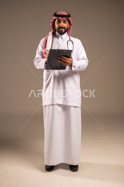 دراسة الحالات المرضية وتقديم الرعاية، بورتريه لطبيب عربي خليجي سعودي يرتدي الثوب التقليدي والشماغ والمعطف الأبيض يمسك بيده ملفات المرضى يقوم بتدوين البيانات ينظر إلى الكاميرا، مهن ووظائف طبية في المملكة العربية السعودية، الجسم بالطول الكامل، خلفية بيج
