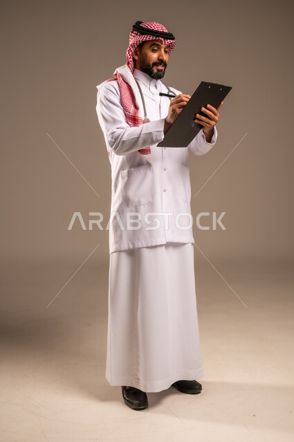 دراسة الحالات المرضية، بورتريه لطبيب عربي خليجي سعودي يرتدي الثوب التقليدي والشماغ والمعطف الأبيض يمسك بيده ملفات المرضى يقوم بتدوين البيانات، مهن ووظائف طبية في المملكة العربية السعودية، تقديم الاستشارات والرعاية الصحية بكفاءة، الجسم بالطول الكامل، خلفية بيج