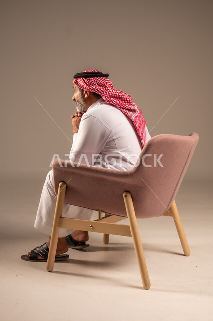التأمل وشرود الذهن، التفكير في شيء هام، بورتريه من الجانب لرجل مسن عربي خليجي سعودي يرتدي الثوب التقليدي والشماغ يجلس على الكرسي يضع يده على ذقنه وينظر الى شيء، خلفية بيج